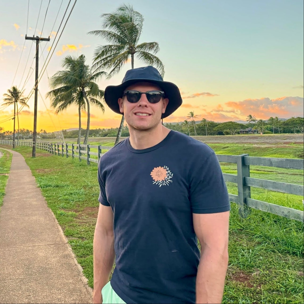 Man wearing a navy blue t-shirt with a logo, sunglasses, and a bucket hat standing on a path with palm trees and a sunset in the background.