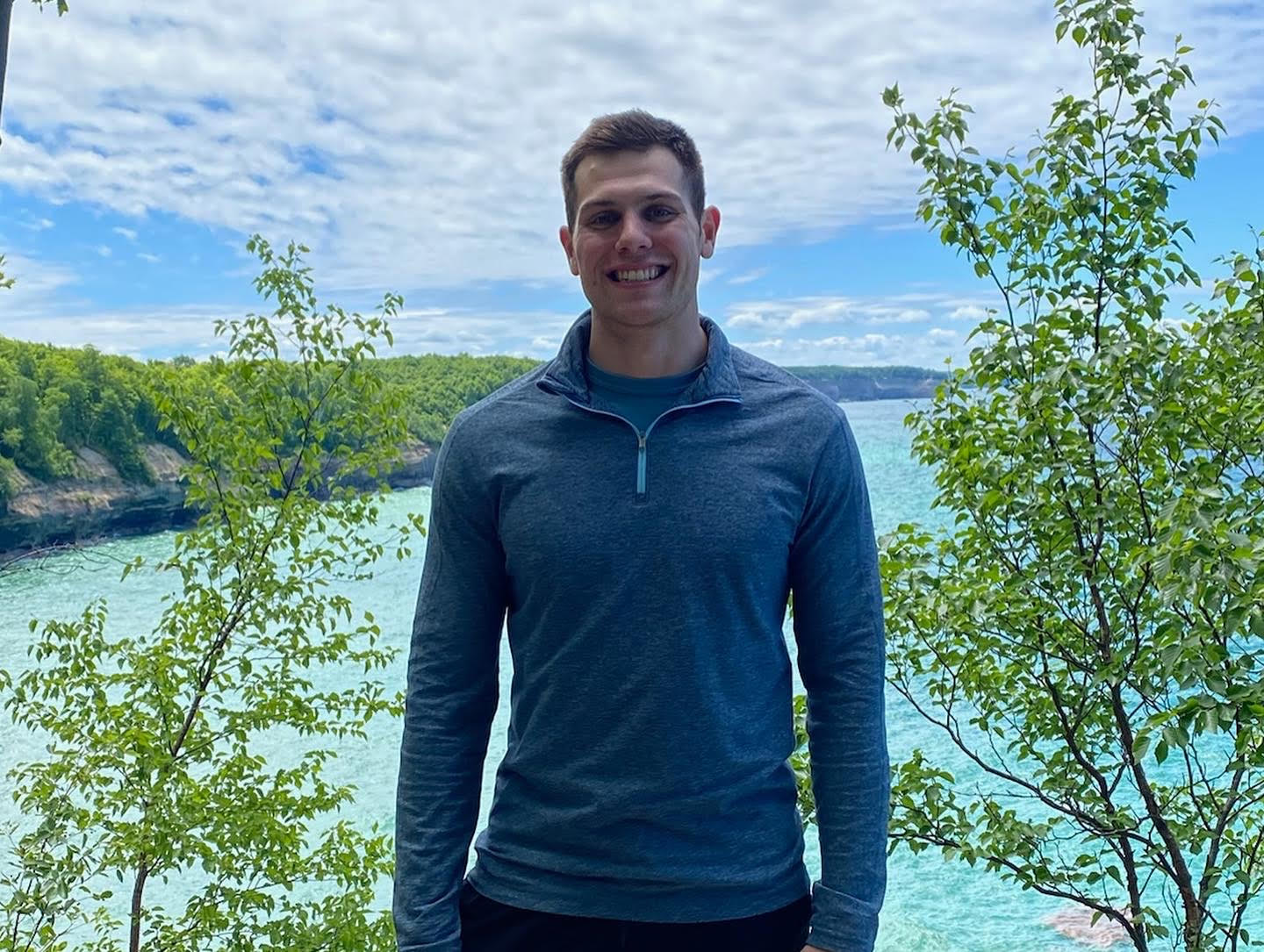 Man in a blue pullover standing in front of a scenic view with trees and water.